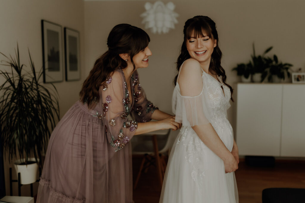 Trauzeugin schnürt das Kleid der Braut am Hochzeitstag beim Getting Ready zu, während sich beide anlachen