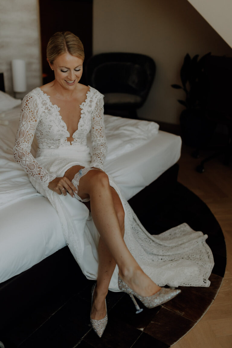 Braut sitzt beim Getting Ready an ihrem Hochzeitstag auf einem Bett und zieht sich ein Strumpfband an