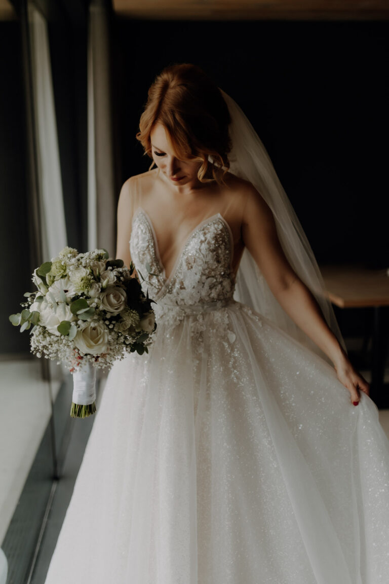 Braut steht mit Brautstrauß und in ihrem Hochzeitskleid beim Getting Ready an einem Fenster