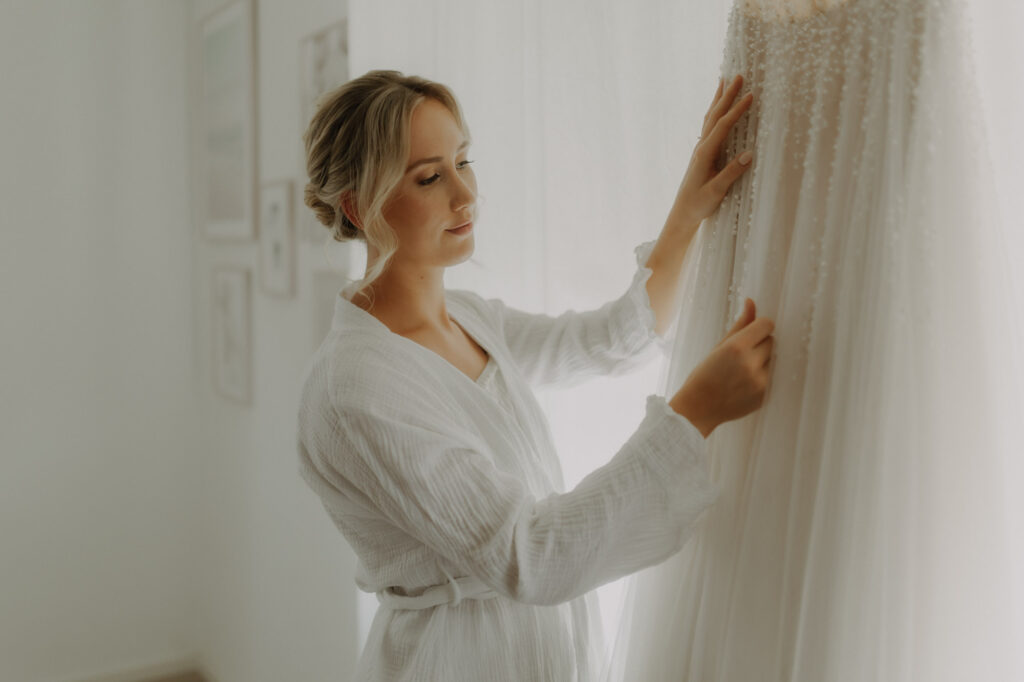 Braut schaut sich ihr Hochzeitskleid am Hochzeitstag beim Getting Ready verliebt an